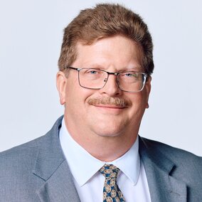 Phllip Myers-Man in a gray suit and patterned tie against a light background, face blurred.